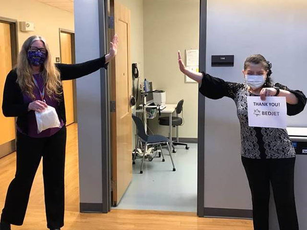 Two healthcare workers wearing masks stand 6+ feet apart and pretend to high five, one holds a sign that says