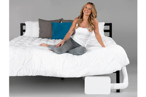 a smiling young blonde woman sitting at the foot of the bed where a BedJet unit is installed