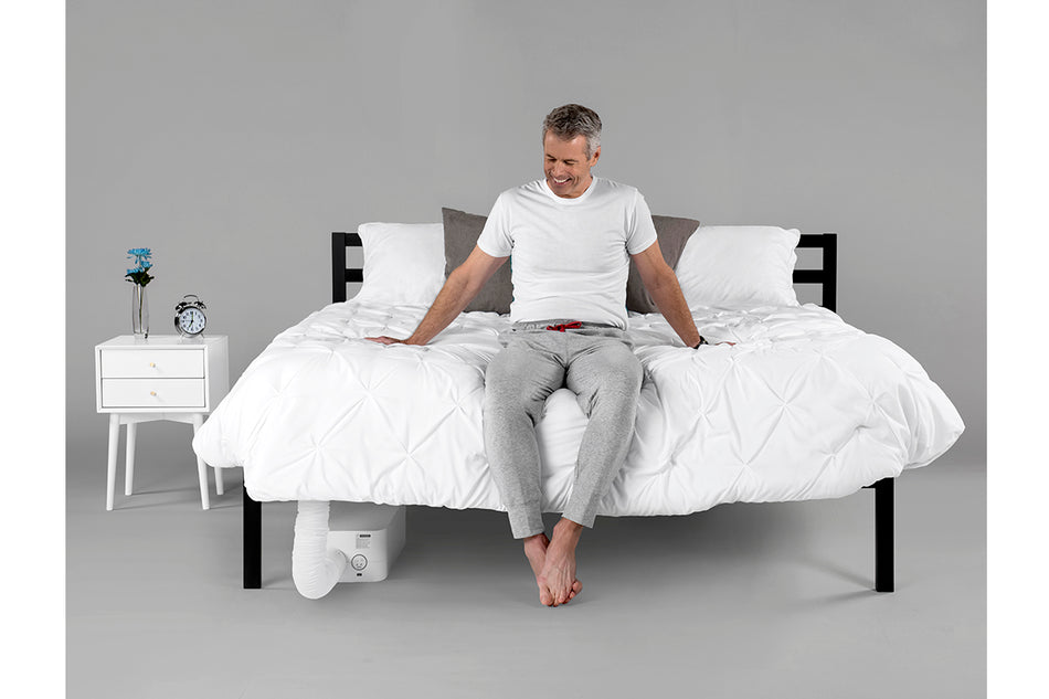  an older man sitting on a bed smiling at a BedJet unit installed at the foot of the bed