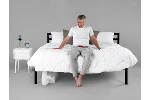 a smiling older man sitting on the edge of a bed looking at a BedJet unit installed underneath