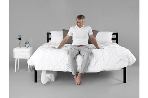 a smiling older man sitting on the edge of a bed looking at a BedJet unit installed underneath