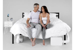 a couple smiling at each other while sitting at the foot of a bed with 2 BedJet units installed